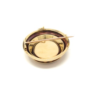 May include: A gold-toned brooch with a round, beveled design. The brooch has a pin on the back and a decorative edge.