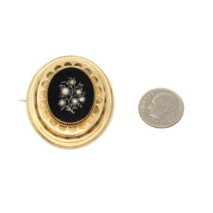 May include: A gold brooch with a black enamel oval surrounded by a gold filigree design. The oval features a floral design with white seed pearls.