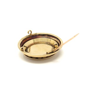 May include: A gold-toned brooch with a round, beveled design and a pin on the back. The brooch has a decorative border and a smooth, circular center.