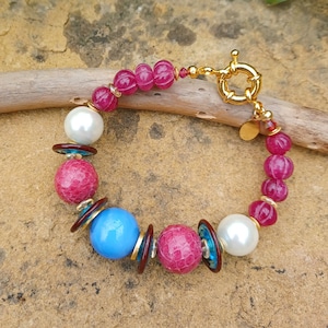 Op de afbeelding: Een roze, blauwe en witte kralen armband met een gouden sluiting. De armband heeft verschillende ronde kralen, waaronder sommige met een getextureerd oppervlak.