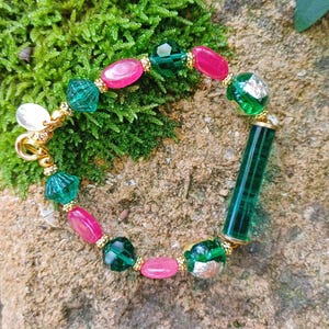 May include: A green and pink beaded bracelet with a gold clasp and a long green glass bead. The bracelet is on a brown and green mossy surface.
