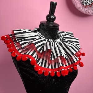 May include: A ruffled neck collar featuring a black and white vertical striped fabric pattern. The collar is layered with a red mesh tulle trim and finished with a decorative red pom-pom fringe along the outer edge, creating a full, pleated look.