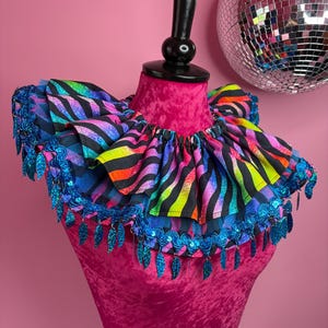 May include: A ruffled neck collar featuring a vibrant rainbow zebra print pattern. The collar is layered with black mesh fabric and finished with a decorative trim of shimmering blue sequined leaf-shaped dangles along the outer edge.