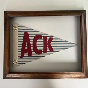 May include: Framed pennant flag with the word "ACK" in red letters. The flag has blue and white stripes and is mounted in a dark brown wooden frame. The pennant is a nautical-themed decorative item.