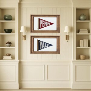 May include: Two framed pennants, one red with "PENN" and the other blue with "YALE", hang on a cream-colored wall. The pennants are in wooden frames. The wall has built-in shelves and sconces.