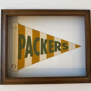 May include: A framed pennant with a yellow and white striped background and the word "PACKERS" in green letters.