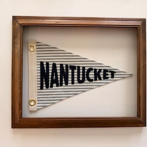 May include: A framed pennant flag with the word "NANTUCKET" in navy blue letters. The flag has a blue and white striped pattern and is framed in a dark brown wooden frame. The flag is attached to a beige fabric strip with gold grommets.