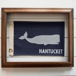 May include: A rectangular navy blue flag featuring a light gray whale silhouette and the white text NANTUCKET. The flag has a white canvas header with two brass grommets on the left side and is displayed inside a dark brown wooden frame.