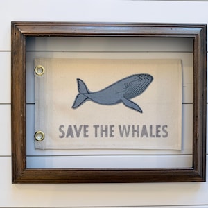 May include: A rectangular off-white canvas flag featuring a blue whale illustration and the text "SAVE THE WHALES" in blue capital letters. The flag has two brass grommets on the left side and is displayed inside a dark brown wooden frame.