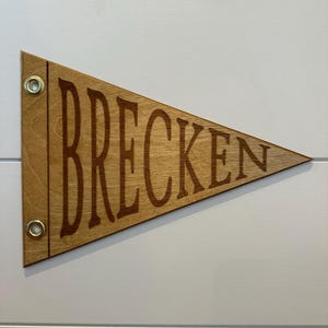May include: A wooden pennant with the word "BRECKEN" engraved in brown lettering. The pennant is light brown and has two brass grommets at the top.