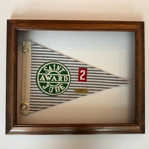 May include: Framed pennant with a blue and white striped pattern. The pennant features a green circular patch with the words "Saint Award Jude" and the number "2" in red. The pennant is framed in a dark brown wooden frame.