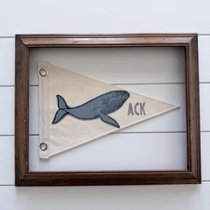 May include: A framed pennant with a whale design. The pennant is cream-colored and features a gray whale applique. The word "ACK" is printed in gray on the pennant. The frame is made of dark brown wood.