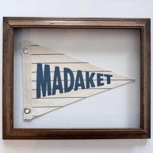 May include: A framed pennant flag with the word "MADAKET" in navy blue block letters. The flag is white with blue horizontal stripes and is framed in a dark brown wooden frame.
