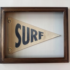 May include: A framed pennant with the word "SURF" in navy blue lettering. The pennant is a light beige color and is framed in a dark brown wooden frame. The pennant is a vintage style and is a decorative item.