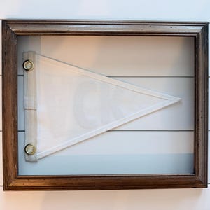 May include: A framed pennant with a white triangular flag. The flag is mounted inside a dark brown wooden frame. The pennant is white with a white border and is attached to the frame with metal grommets.
