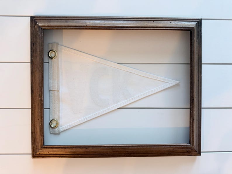 May include: A framed pennant with a white triangular flag. The flag is mounted inside a dark brown wooden frame. The pennant is white with a white border and is attached to the frame with metal grommets.