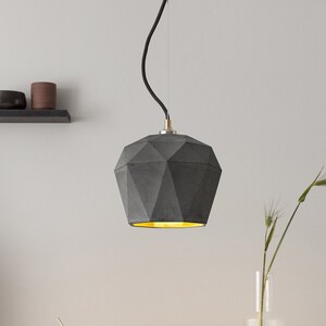 Op de afbeelding: Een grijze betonnen hanglamp met een geometrisch ontwerp hangt aan een zwart snoer. De lamp wordt verlicht met een warm geel licht.