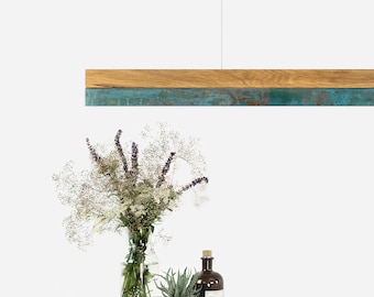 May include: A modern pendant light fixture with a rectangular wooden frame and a copper-toned metal panel. The light is hanging over a white table with a vase of flowers, a potted succulent, and a bottle.