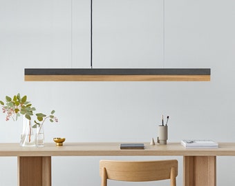 May include: A modern, minimalist pendant light fixture with a rectangular wood and black metal shade. The light is hanging over a wooden table with a vase of flowers and a book.