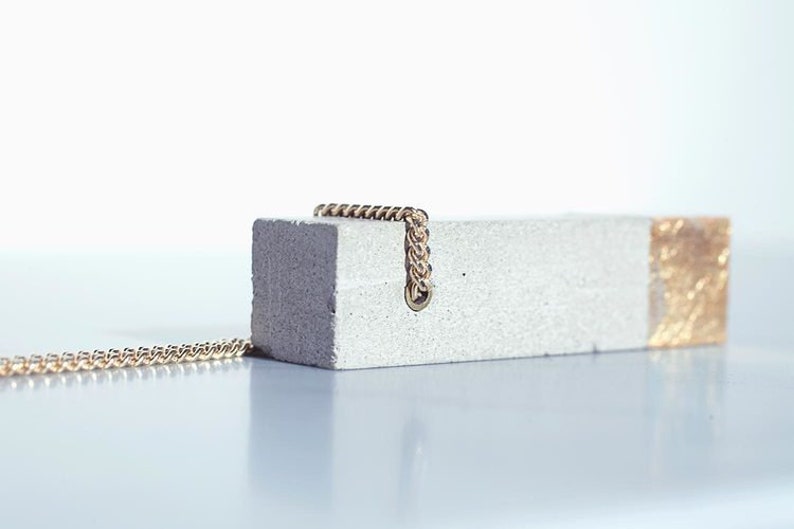May include: A concrete pendant necklace with a gold accent and a gold chain. The pendant is rectangular and has a rough texture.