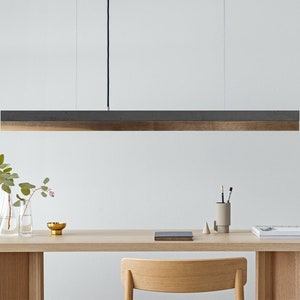 May include: A modern, minimalist pendant light fixture with a rectangular, wood and concrete shade. The light is hanging over a wooden table with a vase of flowers and a notebook.