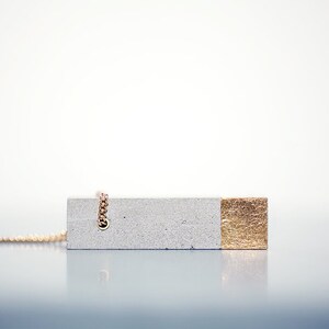 May include: A gray concrete pendant necklace with a gold accent on one side. The necklace is made of a chain with a small loop at the top.