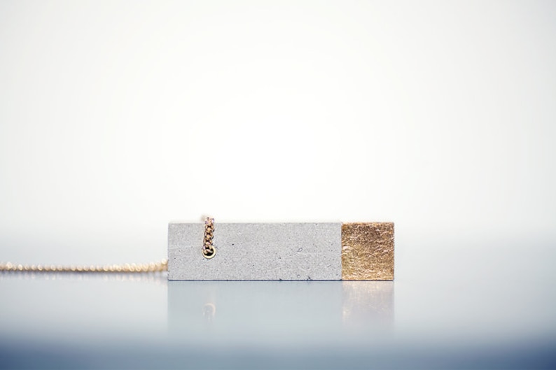 May include: A gray concrete pendant necklace with a gold accent on one side. The necklace is made of a chain with a small loop at the top.