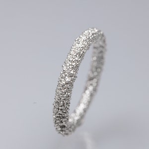 Könnte beinhalten: Ein silberner Ring mit strukturierter Oberfläche, der mit kleinen, funkelnden Details bedeckt zu sein scheint. Der Ring ist vor einem schlichten, hellen Hintergrund platziert, der sein Design hervorhebt. Die Form des Rings ist eine durchgehende Schleife, die ein Band suggeriert.