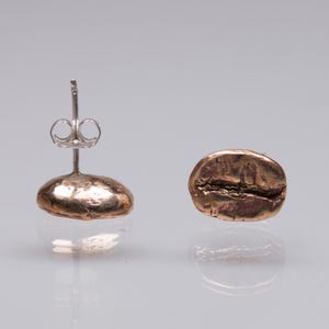 Puede incluir: Un par de pendientes con forma de grano de café. Uno es un pendiente de botón con un poste plateado, y el otro es un grano suelto. Los pendientes son de color bronce y tienen una superficie texturizada que refleja la luz.
