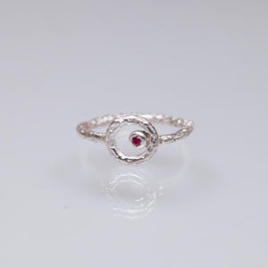 May include: A silver ring with a textured band and a circular design. The ring features a small, round, red gemstone set in the center of the circle. The ring is displayed on a white surface.