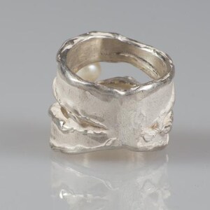 May include: A silver ring with a textured, organic design. The ring has a pearl set into the band.