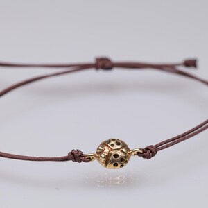 Könnte beinhalten: Braunes Kordelarmband mit einem goldfarbenen, kugelförmigen Anhänger mit kleinen Ausschnitten. Das Armband ist mit Schiebeknoten verstellbar, vor weißem Hintergrund.