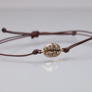 Könnte beinhalten: Ein braunes Kordelarmband mit einem goldfarbenen Kaffeebohnen-Anhänger.