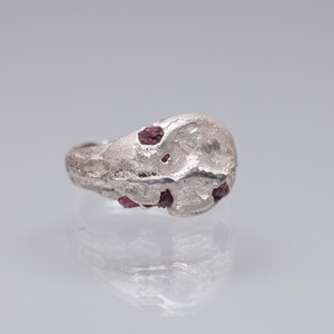 Puede incluir: Un anillo de plata con una superficie texturizada y varias piedras preciosas rojas incrustadas. El anillo tiene una forma orgánica e irregular, las piedras son visibles en la parte superior y los lados.