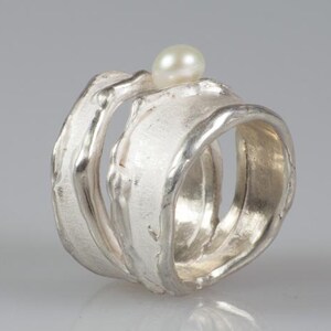 May include: A silver ring with a textured, organic design. The ring is made of two bands that are intertwined. A small white pearl sits on top of the ring.