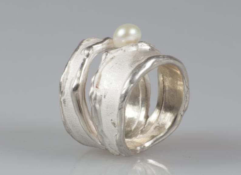 May include: A silver ring with a textured, organic design. The ring is made of two bands that are intertwined. A small white pearl sits on top of the ring.