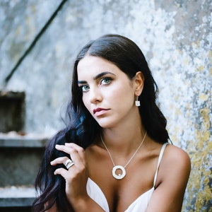 May include: A woman with long dark hair wearing a white sleeveless top and silver jewelry, including a necklace with a large circular pendant, earrings, and a ring. She is sitting on a set of stairs with a textured concrete wall behind her.