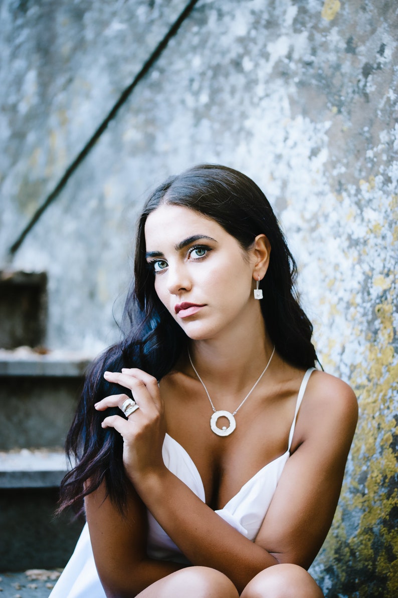 May include: A woman with long dark hair wearing a white sleeveless top and silver jewelry, including a necklace with a large circular pendant, earrings, and a ring. She is sitting on a set of stairs with a textured concrete wall behind her.