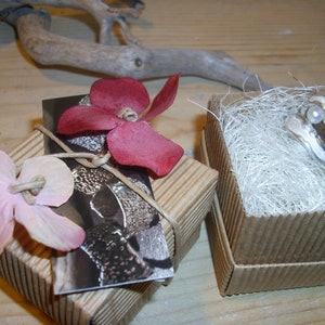 May include: Two small cardboard boxes with decorative flowers on top. One box contains a silver ring with a pearl, the other box contains a silver ring with a textured surface.