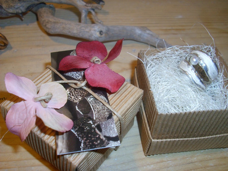 May include: Two small cardboard boxes with decorative flowers on top. One box contains a silver ring with a pearl, the other box contains a silver ring with a textured surface.