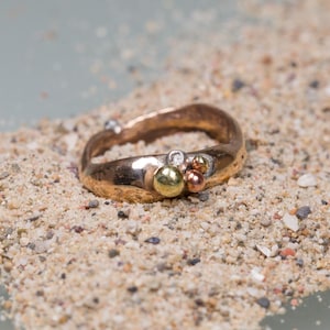 Puede incluir: Un anillo de oro con una superficie texturizada y tres pequeñas piedras redondas engastadas en la banda. Las piedras son una mezcla de oro, verde y blanco.