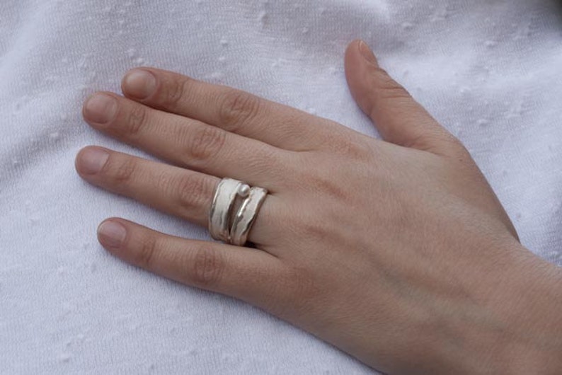 May include: A silver ring with a white enamel design. The ring is worn on the middle finger of a hand.