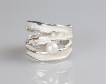 May include: A silver ring with a textured, layered design. The ring features a large, round, white pearl at its center. The ring's design is organic and modern, with a reflective surface.