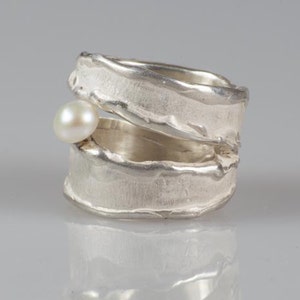 May include: A silver ring with a textured, organic design. The ring is open and has a small pearl set into the side.