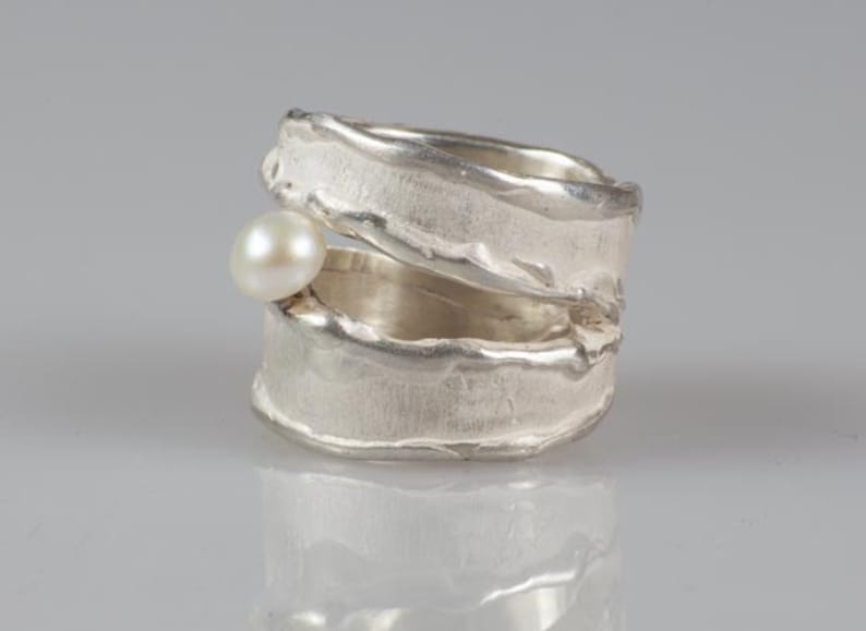 May include: A silver ring with a textured, organic design. The ring is open and has a small pearl set into the side.