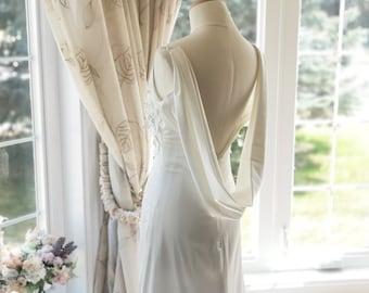 May include: A white wedding dress with a draped cowl back detail is displayed on a mannequin. The dress has a simple, elegant silhouette and is set against a window with floral curtains.