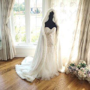 May include: A white faux fur bridal cape with a hood, draped over a white wedding dress with lace detailing. The cape is shown on a mannequin in a room with white curtains and a wooden floor.