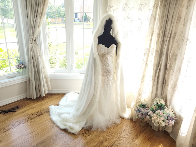 May include: A white faux fur bridal cape with a hood, draped over a white wedding dress with lace detailing. The cape is shown on a mannequin in a room with white curtains and a wooden floor.