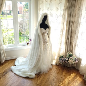 A white hooded cape made of luxurious velvet with delicate white trim. The cape features a small train, adding an elegant touch. Ideal for winter weddings, providing warmth and a glamorous feel.