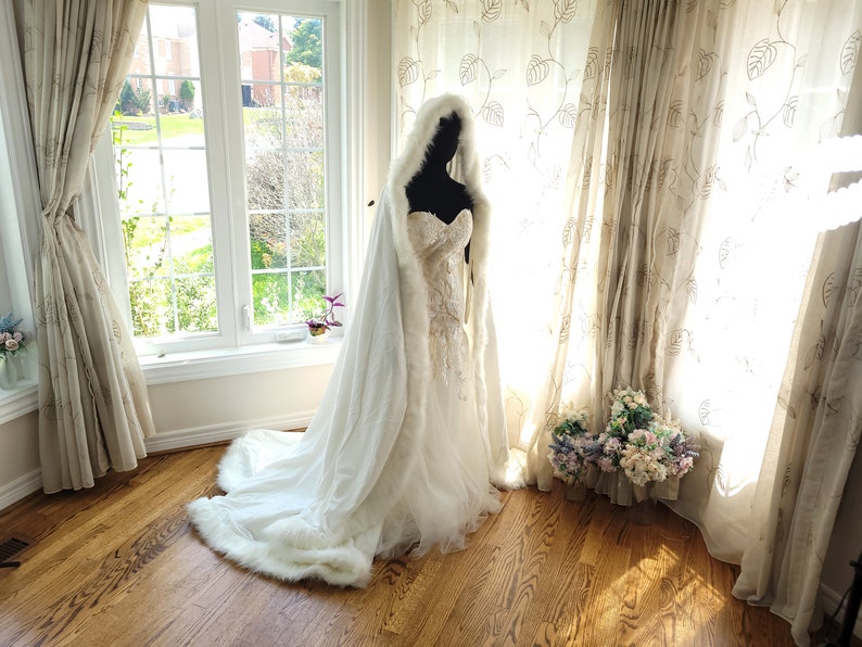 A white hooded cape made of luxurious velvet with delicate white trim. The cape features a small train, adding an elegant touch. Ideal for winter weddings, providing warmth and a glamorous feel.